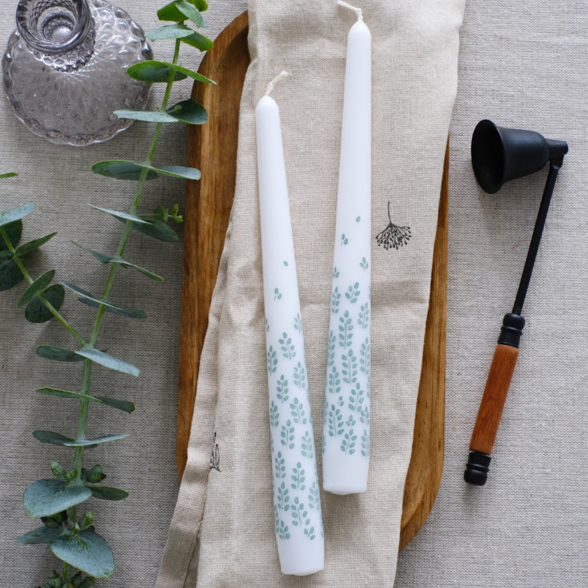 Two tapered dinner candles, white, with sage green leafy detail hand painted on to them. The candles are laid on a organic cotton cloth with a glass candlestick beside them, some eucalyptus and a candle snuffer.