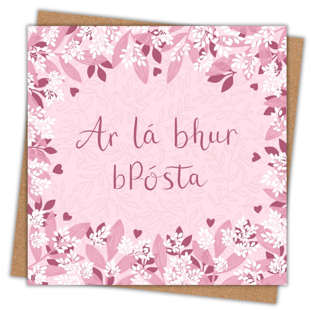 Irish botanical wedding card with the words "Ar lá bhur bpósta" written in dark pink hand writing, surrounded by clusters of light and dark pink leaves and small white leaves. The background of the card is a very light pink. Sitting behind the card is a recycled brown Kraft envelope.