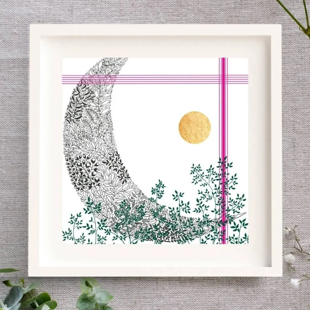 Framed art print on a grey/brown linen cloth with eucalyptus and baby's breath around it. The frame is white. It's square. There is a white mount surround the print. The print is of a black pen illustration of a moon, blended with some green leaves to the bottom of the print. To the top and the right side there are a few bright pink lines and lastly, to the right of centre there is a hand-applied gold leaf circle.