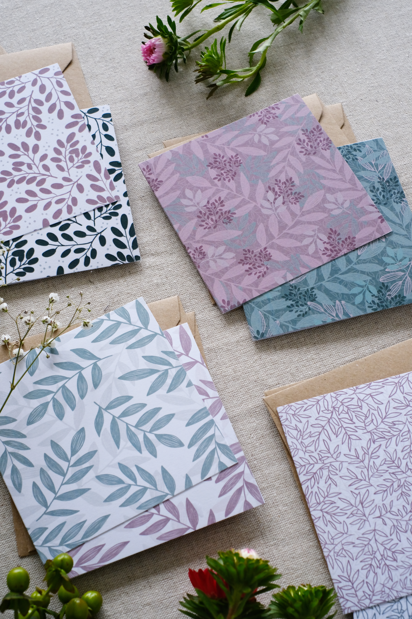 A collection of 8 square botanical-themed note cards, each with their own Kraft brown envelope. The note cards feature intricate leafy patterns in a mixture of green and pink color schemes. The note cards are laid out on a neutral natural fabric surface. Surrounding the card bundles are bunches of greenery, white and pink flowers which go with the natural botanical tone of the sustainably made note cards.