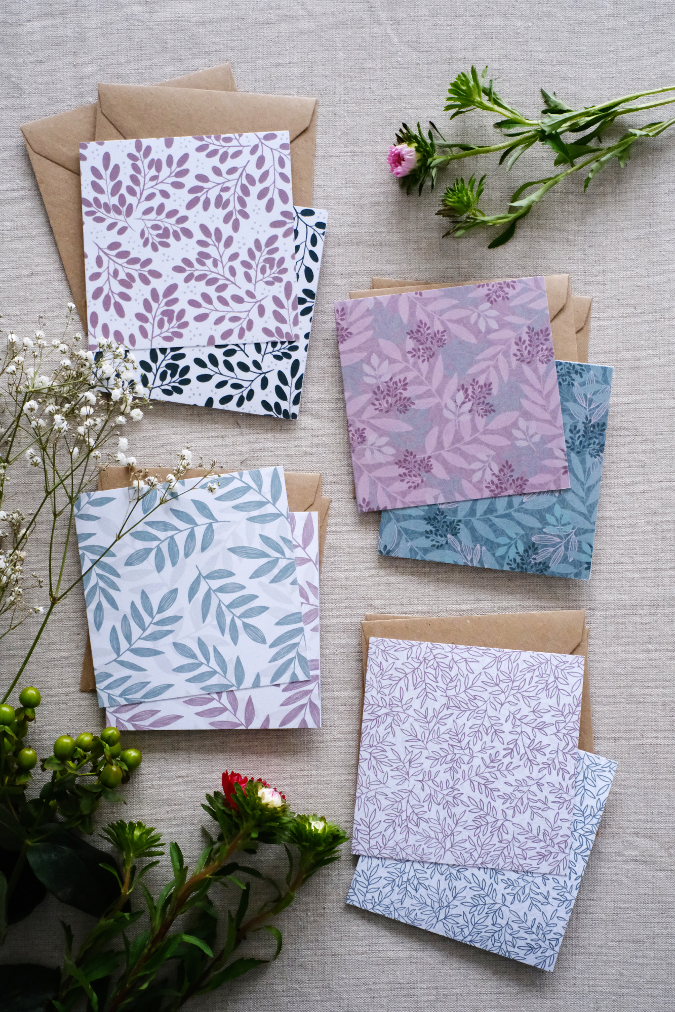 A collection of 8 square botanical-themed note cards, each featuring intricate leafy patterns in green and pink color schemes. The cards are stacked with matching brown kraft envelopes and laid out on a neutral natural fabric surface. Surrounding the card bundles are bunches of greenery, white and pink flowers which go with the natural botanical tone of the note cards.