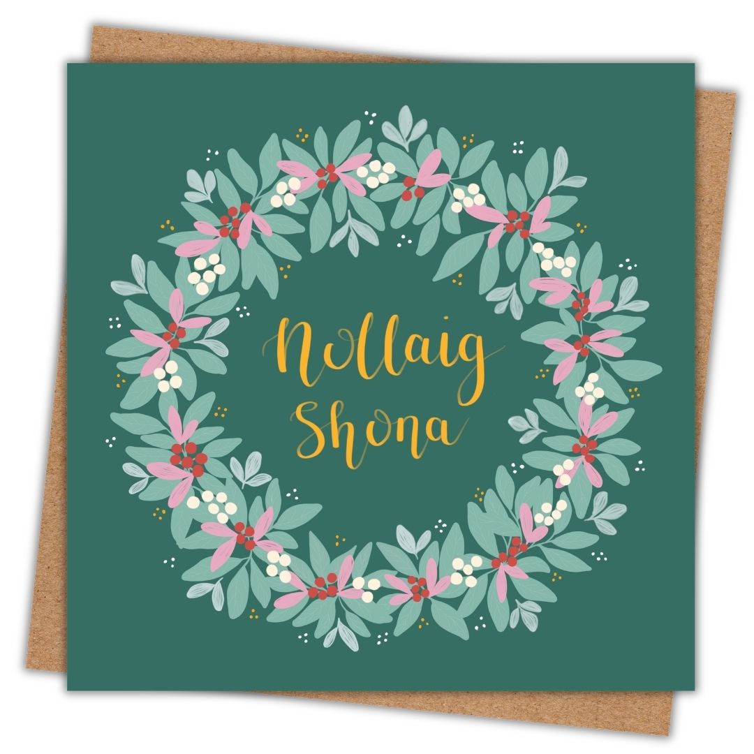 A square greeting card with a dark green background, featuring a decorative wreath made of pale green leaves, pink blossoms, small white berries, and red accents, with the words 'Nollaig Shona' in gold script. The card is shown slightly offset over a brown kraft envelope on a white background.