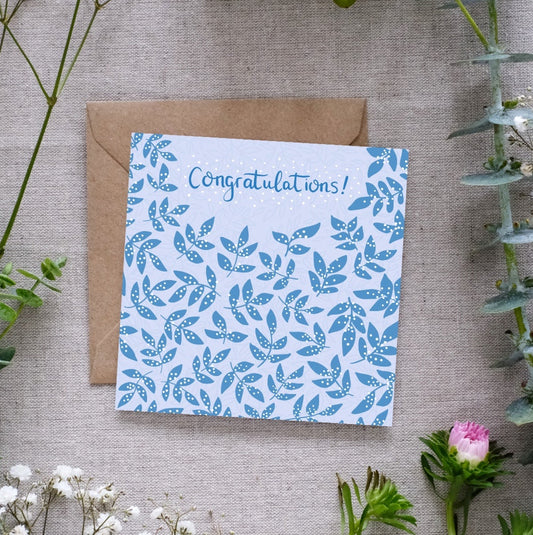 Image shows a congratulations card. The word congratulations is written in dark blue handwriting. In the same dark blue there are clusters of leaves, covered in a white dots which are reminiscent of confetti. The card is sitting on a brown Kraft envelope. Both are laid out on a linen cloth surrounded by light delicate pretty flowers and leaves.