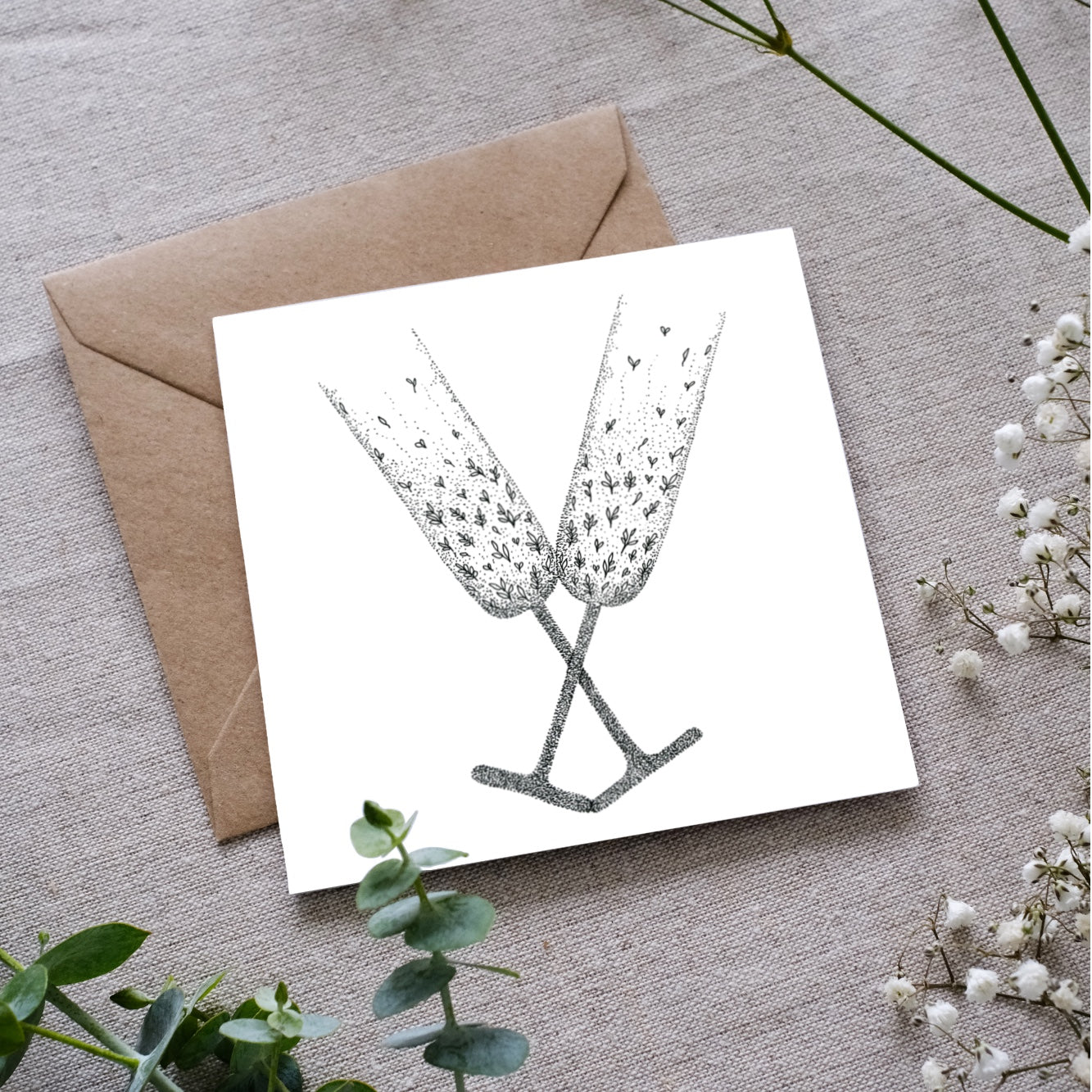 Minimalist black and white card showing two detailed champagne glasses clinking, styled on textured fabric with greenery nearby