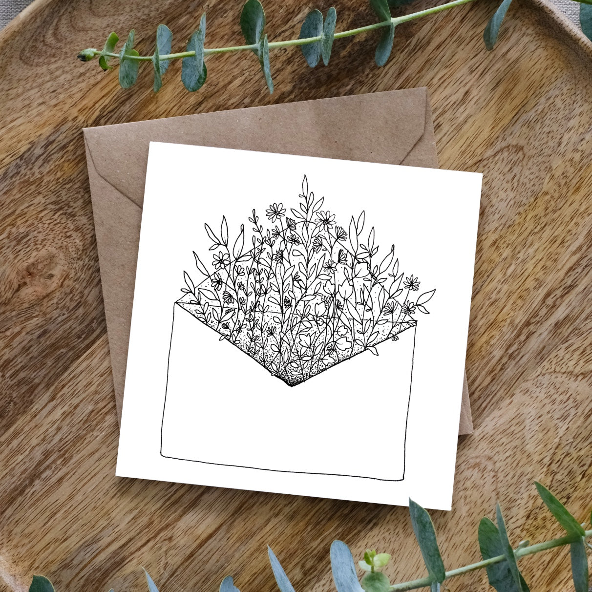 Black and white line drawing of an open envelope overflowing with detailed floral patterns on wooden surface with eucalyptus leaves