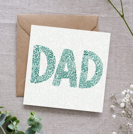 image shows a greeting card with a soft leafy pattern in the background. In the foreground the word DAD is written using intricate leafy details in a beautiful deep green. The card is laid on a brown Kraft envelope. In the background there is a linen cloth and delicate botanical elements.