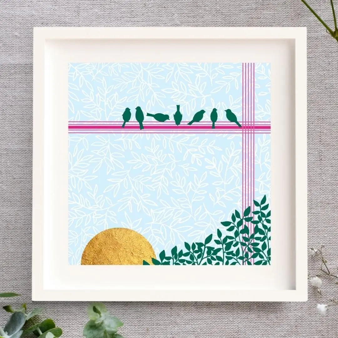 Framed artwork with a light and bright baby blue background with a leaf pattern. The bottom right corner is filled with green leaves. There are seven little birds perched on a hot pink line above a bright rising sun. The sun is made of gold leaf. The artwork is in a white mount and a white solid wood frame. It is laid on a natural cloth and surrounded by folliage.