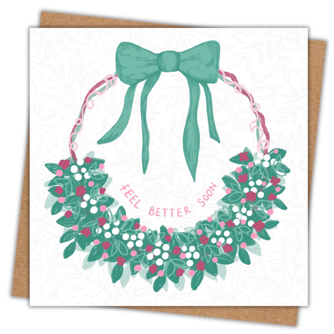 Irish botanical note card which says "Feel Better Soon" in the centre of a floral leafy patterned wreath. The wreath is made of ribbons in green, pink, and dark pink and the leaves cover the bottom half of the wreath. There is a big green bow at the top. Behind the card is a recycled brown Kraft envelope.