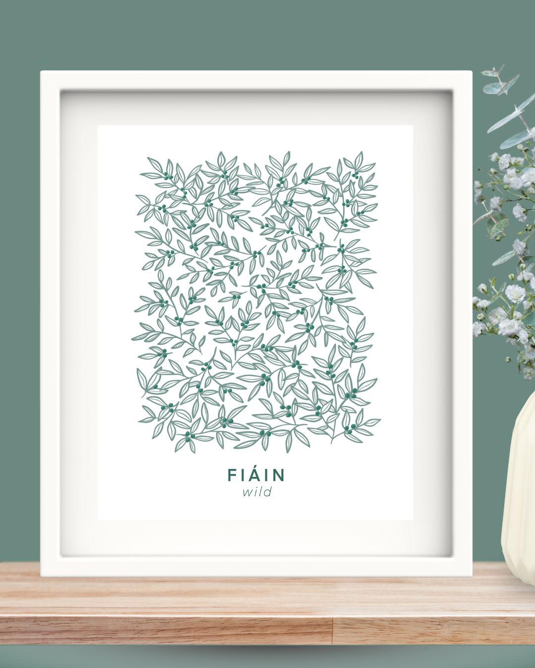 A minimalist framed art print featuring an intricate, hand-drawn botanical pattern of intertwining green leaves and small berries. Below the design, the word 'FIÁIN' is displayed in deep green uppercase letters, with the translation 'wild' written in a delicate, lowercase font beneath it. The artwork is set in a simple Irish made solid wood white frame and white mount. The frame is styled on a wooden shelf, with a deep green wall behind, and a bunch of flowers.
