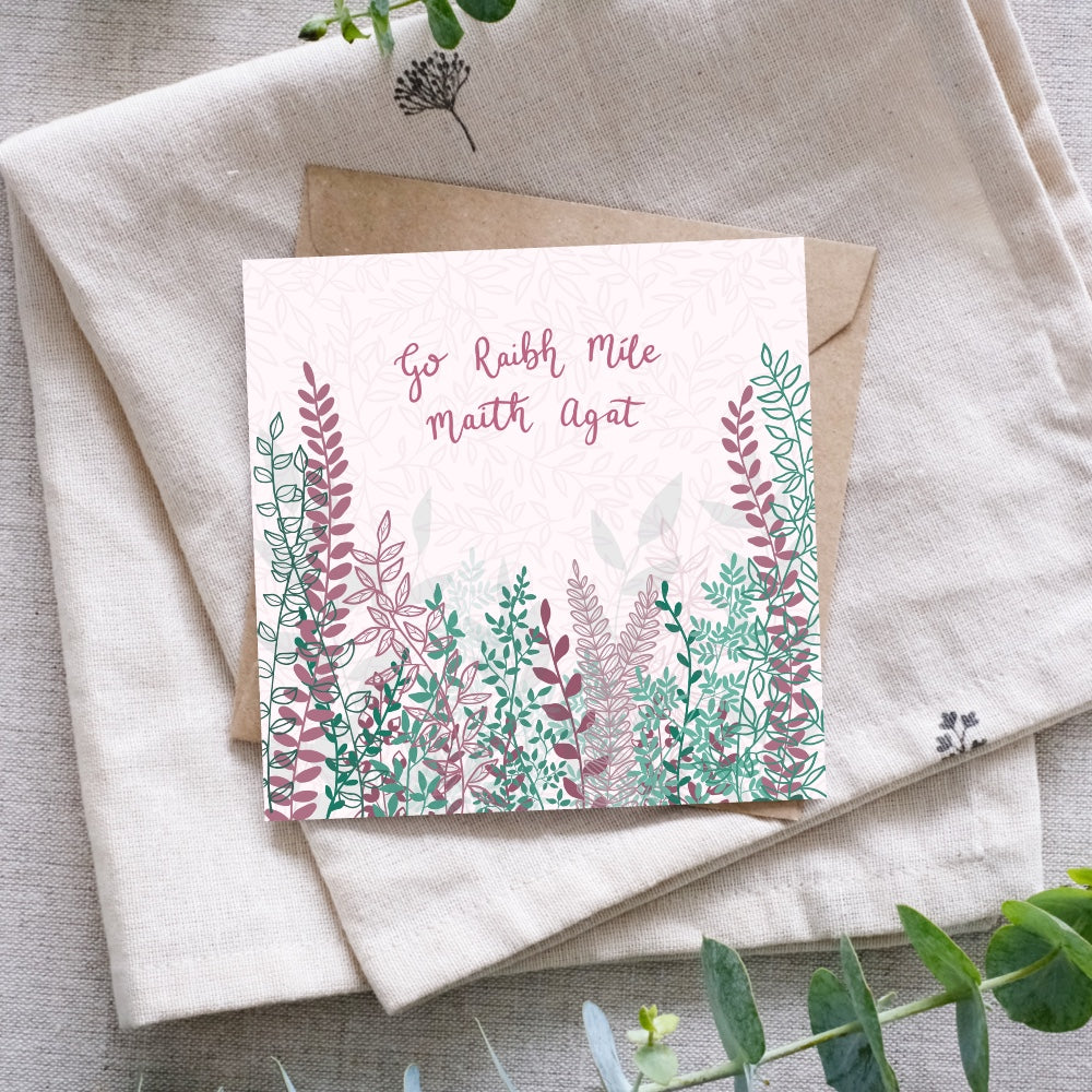 And Irish language thank you card that says "Go Raibh Míle Maith Agat" in Irish along the top of the card. Below the writing are bunches and branches of leaves, and buds in a mixture of dark pink and lots of green tones. Beneath the card is a recycled brown Kraft envelope and they are both sitting on a folded cloth surrounded by foliage for display.