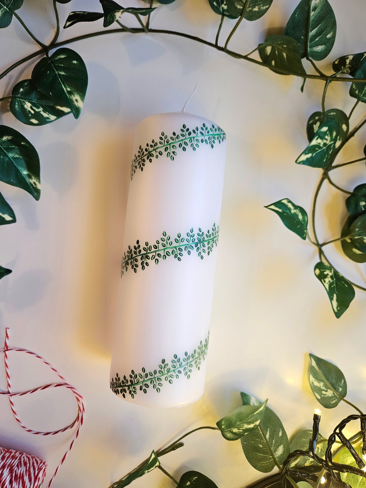 A white pillar candle with a hand-painted leafy design in green, placed on a surface with green leaves and red decorations, likely to be used as a centerpiece for Christmas.
