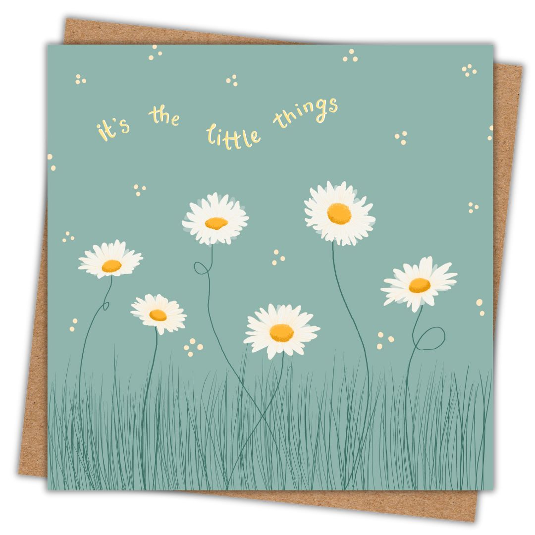 Simple image of a square greeting card with its accompanying Kraft brown envelope. The card has a muted green background, featuring a delicate illustration of white daisies with yellow centers growing in tall grass. Small scattered dots surround the flowers, adding a whimsical touch. Above the floral design, the phrase 'It's the little things' is written in soft yellow, curved text.