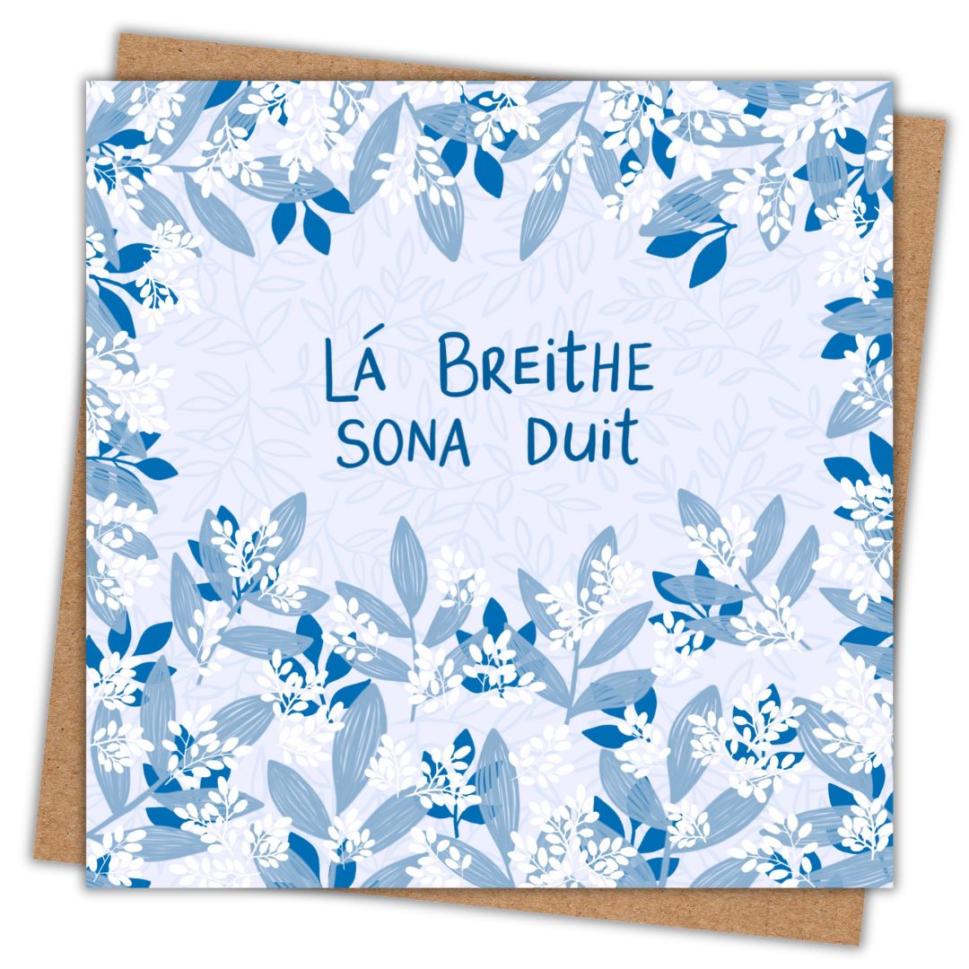 Irish language birthday card with the words Lá Breithe Sona Duit hand written across the centre of the card in dark blue. Surrounding the writing and filling the rest of the card are clusters of light and dark blue leaves of various shapes and sizes and clusters of small white leaves. The background of the birthday card is a soft pale blue. Beneath the card is a recycled brown Kraft envelope.