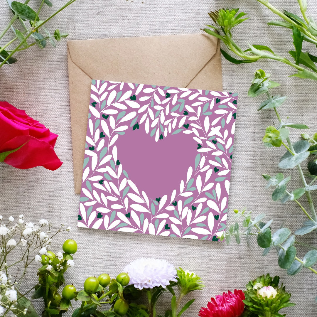 Square greeting card with a muted mauve background, featuring a heart-shaped cutout surrounded by an intricate leafy pattern in white, green, and dark accents. The delicate botanical design creates a romantic and elegant feel. The card is placed on a brown kraft envelope, resting on a textured fabric surface, surrounded by fresh flowers including red roses, white blossoms, and green foliage.