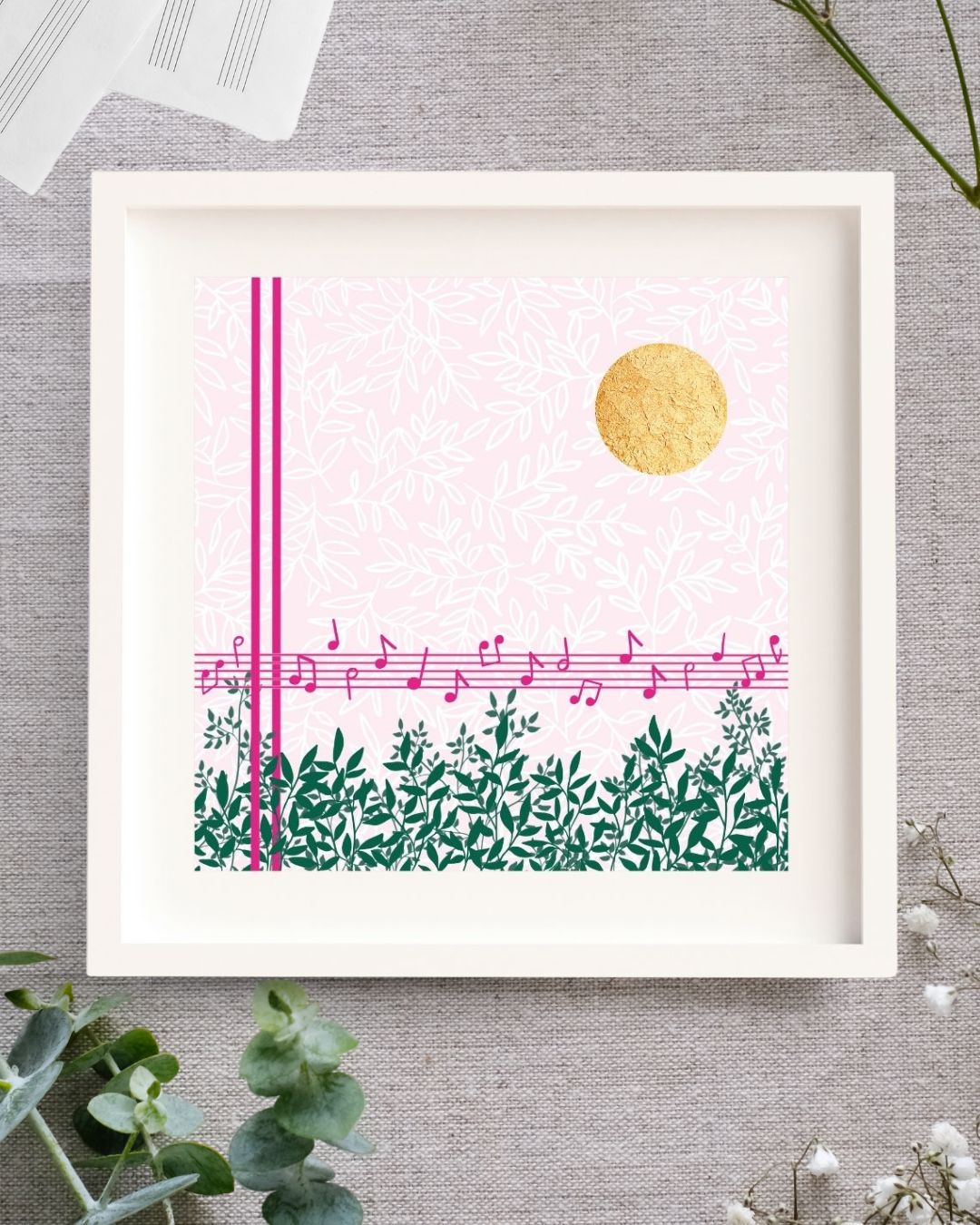 Midday Melody is a square design. The background is a soft light pink with a white leafy pattern. To the fore there are strong pink lines. Two vertical lines, and 5 horizontal lines forming a music stave with a scattering of pink notes on it. Along the bottom there are lots of green branches full of leaves. In the top right corner there is bright circle of gold leaf which has lovely texture to it. The design is framed in a white solid wood Irish made frame. It's displayed on a linen cloth.