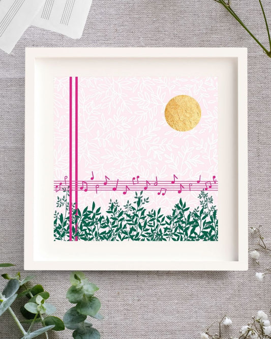 Midday Melody is a square design. The background is a soft light pink with a white leafy pattern. To the fore there are strong pink lines. Two vertical lines, and 5 horizontal lines forming a music stave with a scattering of pink notes on it. Along the bottom there are lots of green branches full of leaves. In the top right corner there is bright circle of gold leaf which has lovely texture to it. The design is framed in a white solid wood Irish made frame. It's displayed on a linen cloth.