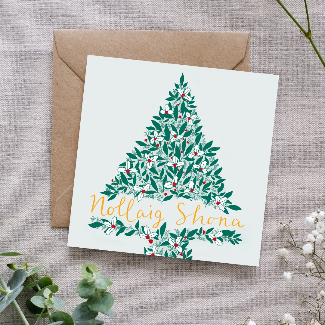 A Christmas tree greeting card with Irish-language text 'Nollaig Shona' and an illustration of a Christmas tree. The tree is made of green leaves, red berries, smaller intricate details white holly leaves. The background of the card is a very subtle soft green. The card is placed on a brown envelope and some decorative foliage in the background.