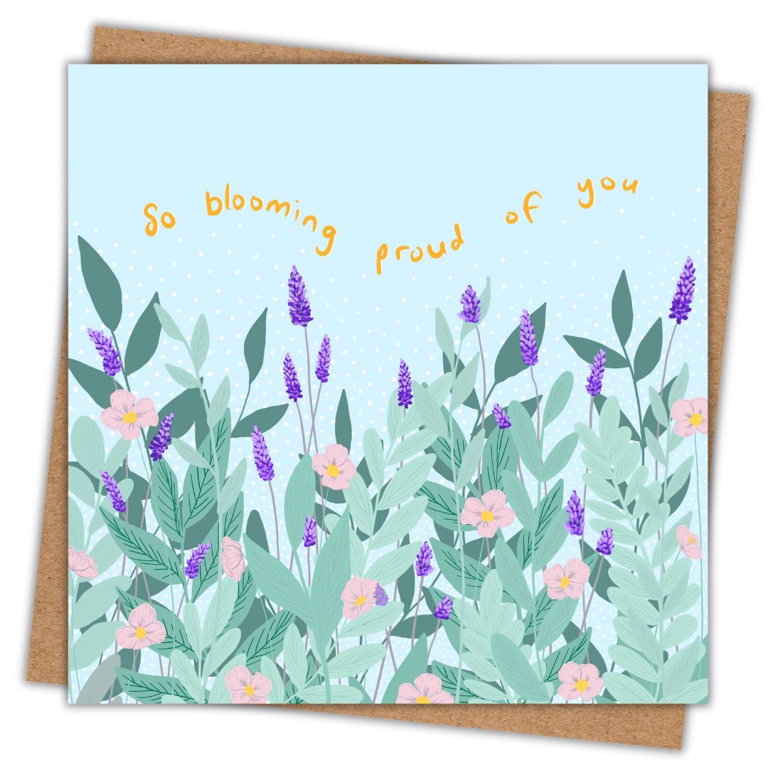 Simple image of a square greeting card with its accompanying Kraft brown envelope behind it. The card has a pastel blue background, featuring an illustrated garden scene with lush green leaves, purple lavender stalks, and delicate pink flowers. Above the floral design, the phrase 'So blooming proud of you' is written in playful, curved yellow text.