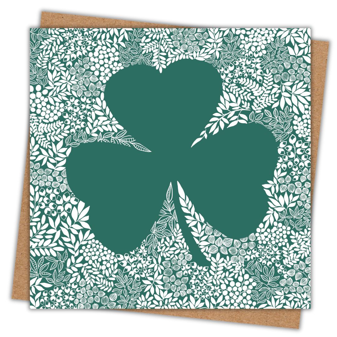 Simple image of a square greeting card with its Kraft brown envelope behind it. The card features a large green shamrock silhouette surrounded by an intricate white floral and leaf pattern. The detailed botanical design contrasts against the deep green background, creating a striking visual effect.
