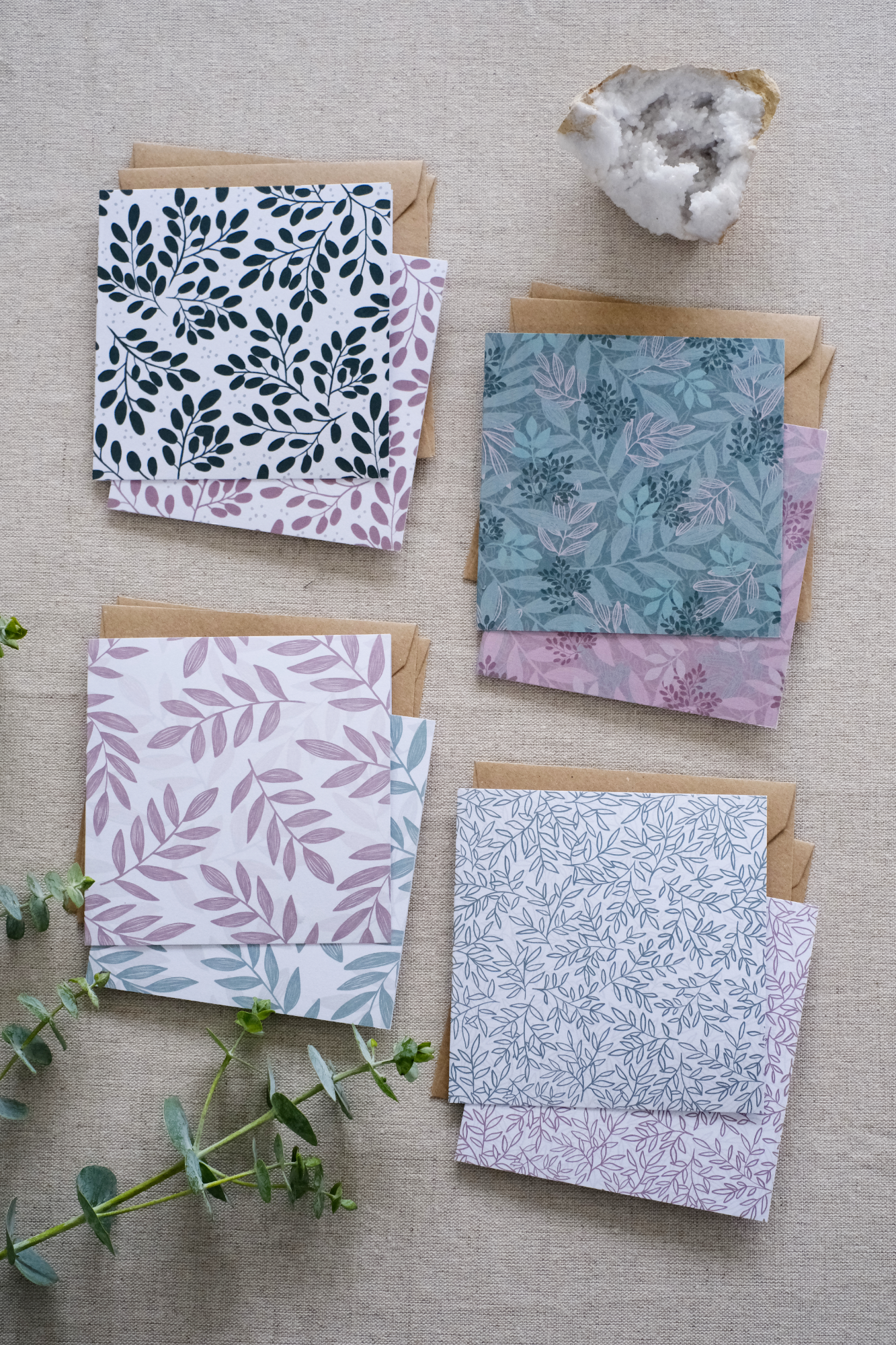 A collection of 8 square botanical-themed note cards, each featuring intricate leafy patterns in green and pink color schemes. The cards are stacked with matching brown kraft envelopes and laid out on a neutral natural fabric surface. Adding more natural tones to the image are sprigs of eucalyptus and greenery and a white crystal.