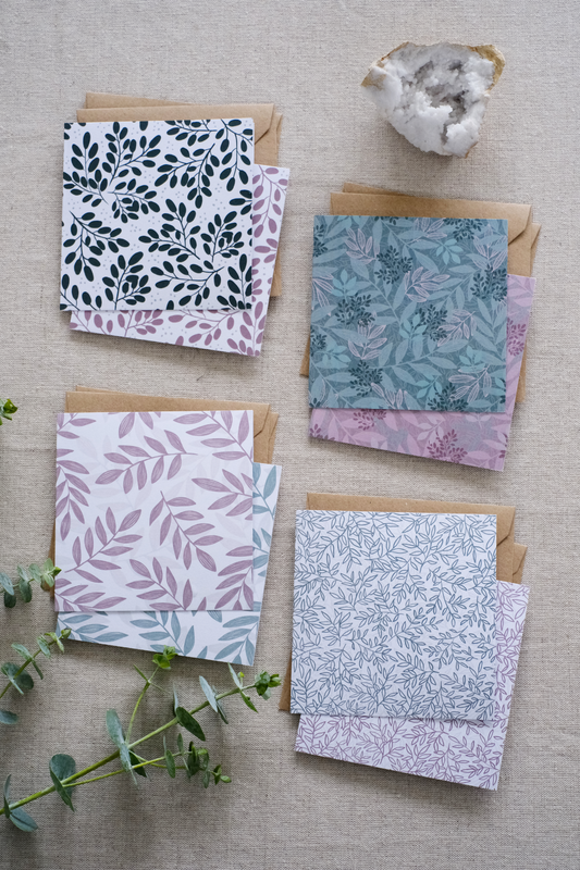 A collection of 8 square botanical-themed note cards, each featuring intricate leafy patterns in green and pink color schemes. The cards are stacked with matching brown kraft envelopes and laid out on a neutral natural fabric surface. Adding more natural tones to the image are sprigs of eucalyptus and greenery and a white crystal.
