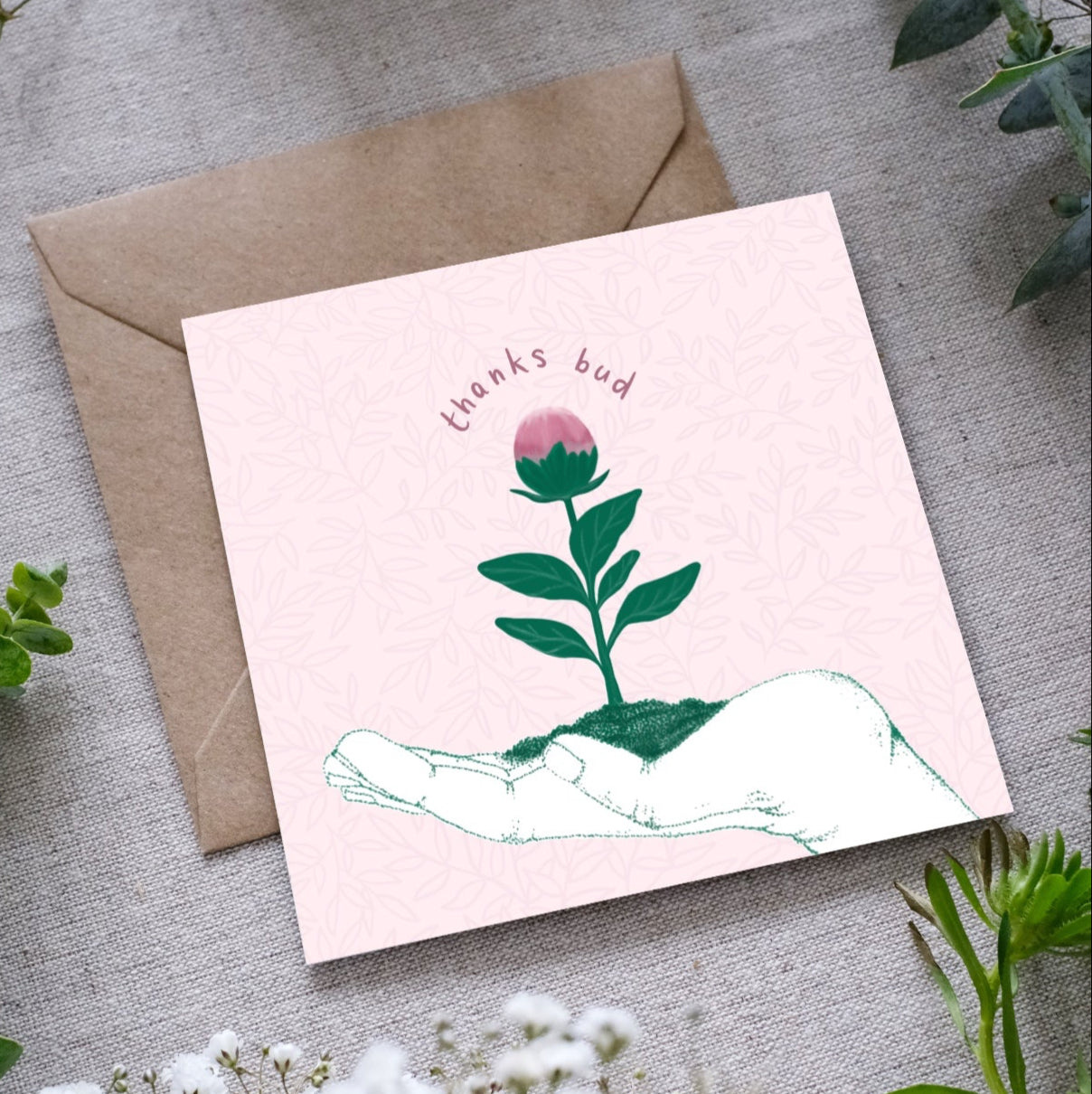 A greeting card with the outline of a hand facing upwards with a little green seedling with a pink bud at the top, sitting in the palm of the hand. The text 'Thanks bud' is written in dusty pink hand writing around the bud of the seedling. The card is accompanied by a brown envelope, displayed on a fabric surface with foliage around.