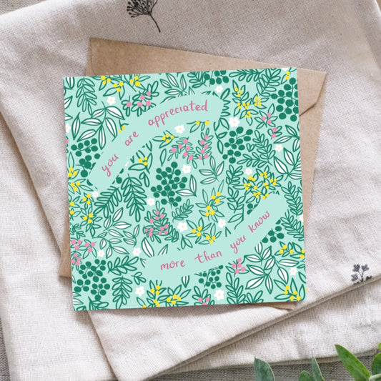 Image shows a pretty thank you card which says You Are Appreciated More Than You Know written in deep pink hand writing. The background of the card is a soft light green with dark green leaves, buds and floral details. There are scatterings of yellow and pink bud clusters as well as pretty daisy heads in between gaps in the leaves. The card is laid on a brown recycled Kraft envelope. Underneath that is a linen cloth for display.