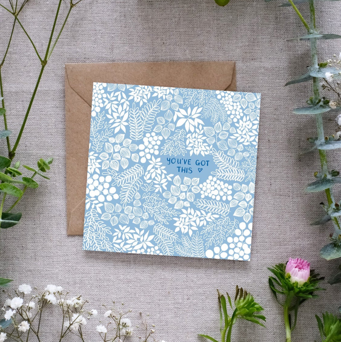 A square greeting card with a soft baby sky blue background and intricate white floral patterns. The words 'You've Got This' are centered in our hand-writing with a tiny heart outline beside it. The card is placed on a textured linen surface, partially resting on a brown kraft envelope. Surrounding the card are sprigs of eucalyptus, baby’s breath, and other greenery, along with a small pink flower.