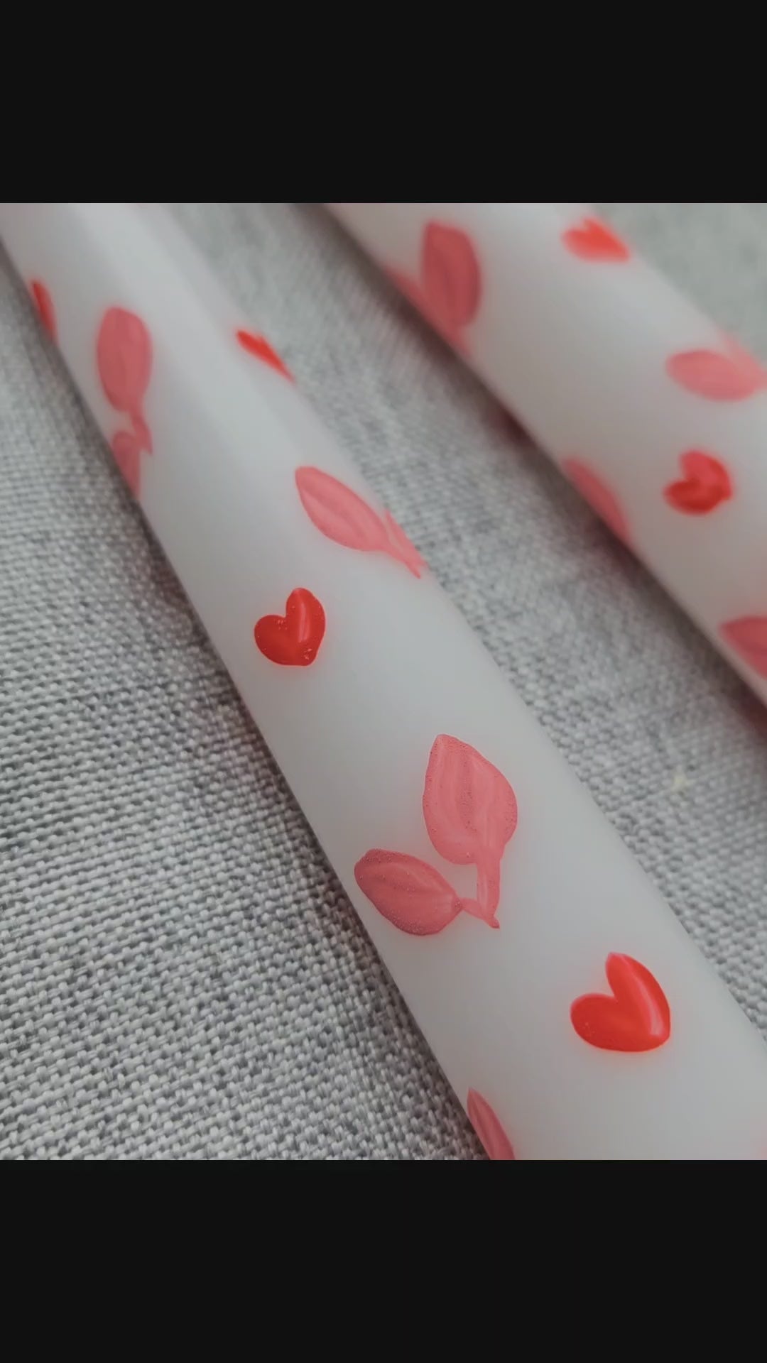 Close-up of white tapered dinner candles where you can see closely the brush strokes on the hand-painted red hearts and pink leaves. There is a grey cloth in the background.
