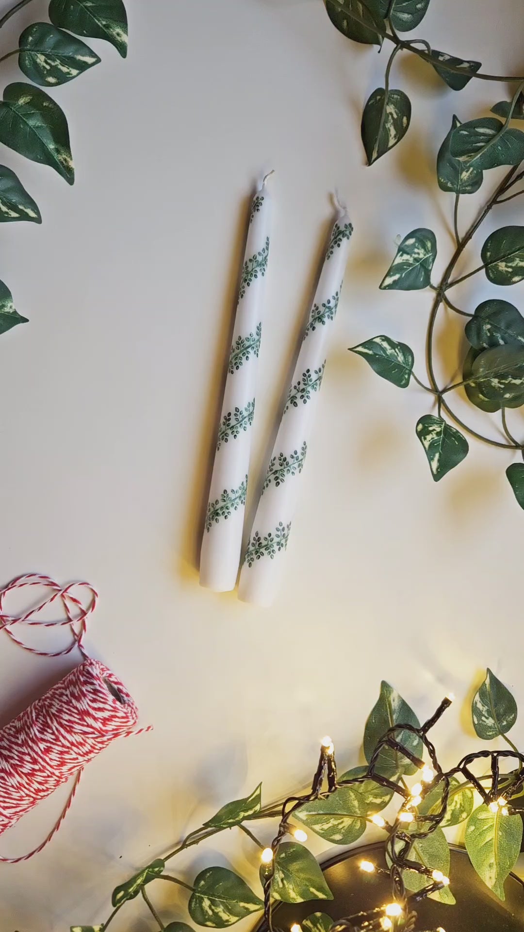 Video of two tapered dinner candles, white, with a green leafy garland design hand painted up around the length of each one in a rich deep green. The video shows the candles up close so you can see the ahdn-painted effect.