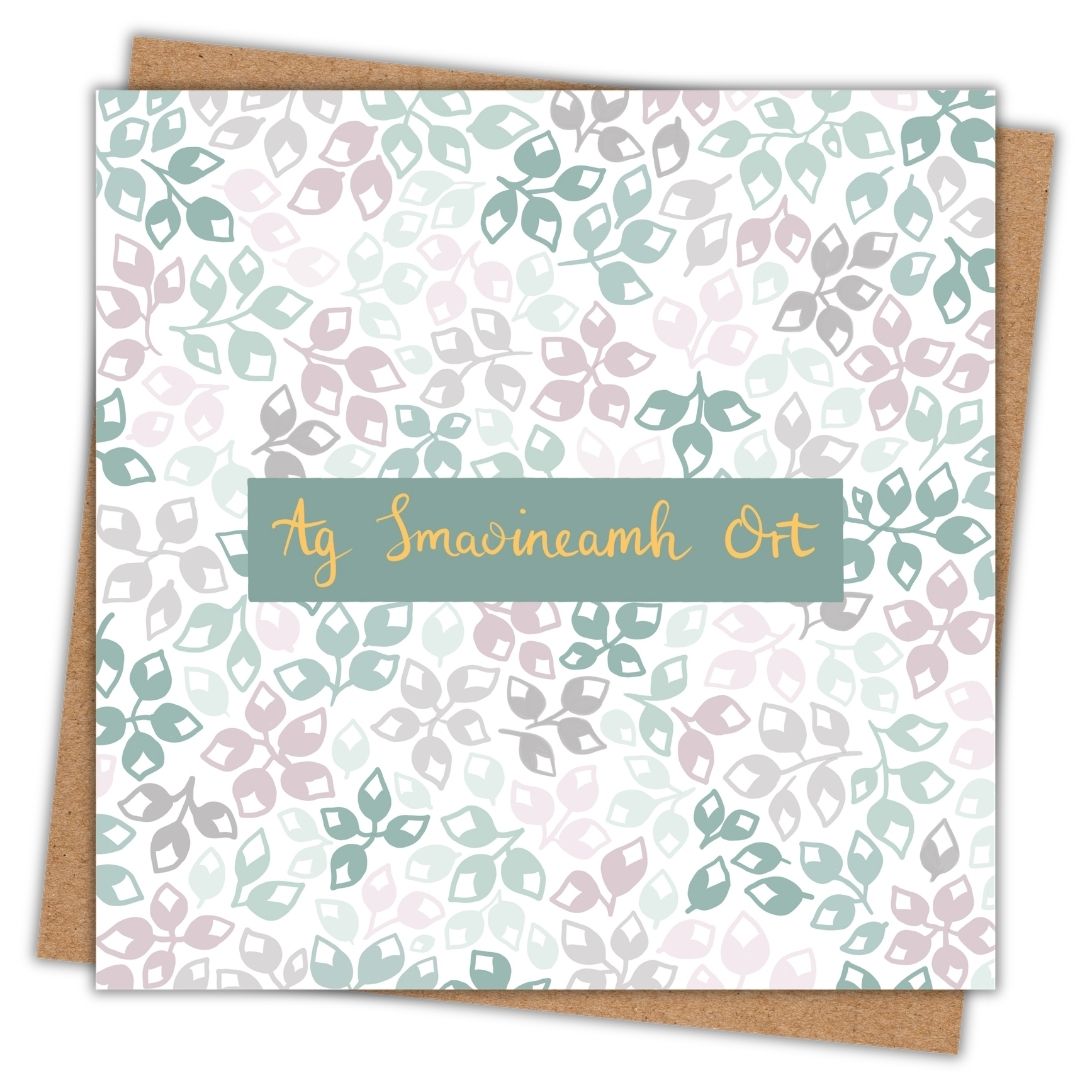 Botanical themed greeting card with simple floral buds of different colours. The colour palette is various shades of green, grey and dusty pink. In the centre of the card are the Gaeilge words "Ag Smaoineamh Ort" in gold writing on a dark green background. Ag Smaoineamh Ort is the Irish language version of the thoughtful phrase "Thinking of You".
