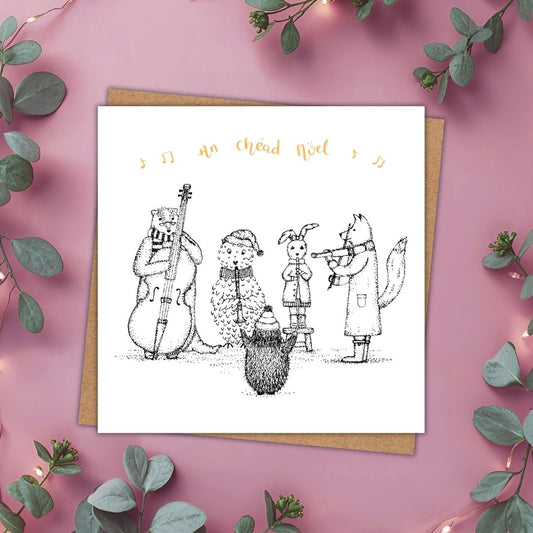 Irish language Christmas card with a group of woodland animals playing music. Above them are some music notes and the words "An chéad Noel" meaning, The First Noel, in gold writing.
