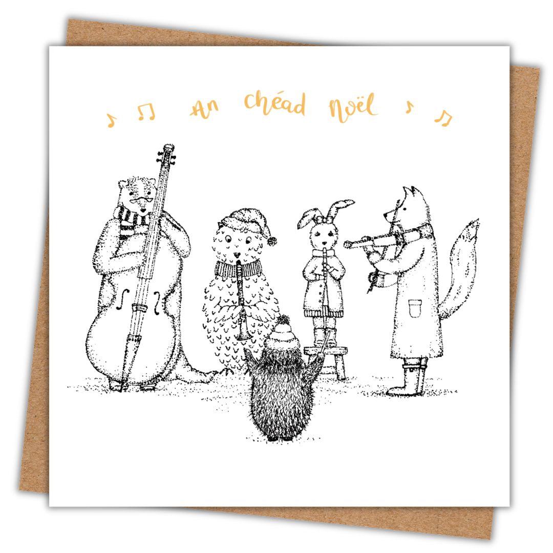 A square white greeting card with a black line-art illustration of winter animals playing music, including a badger, owl, rabbit, fox, and hedgehog. The card has golden handwritten text at the top reading 'An chéad noél' and is shown leaning against a brown kraft envelope on a white background.