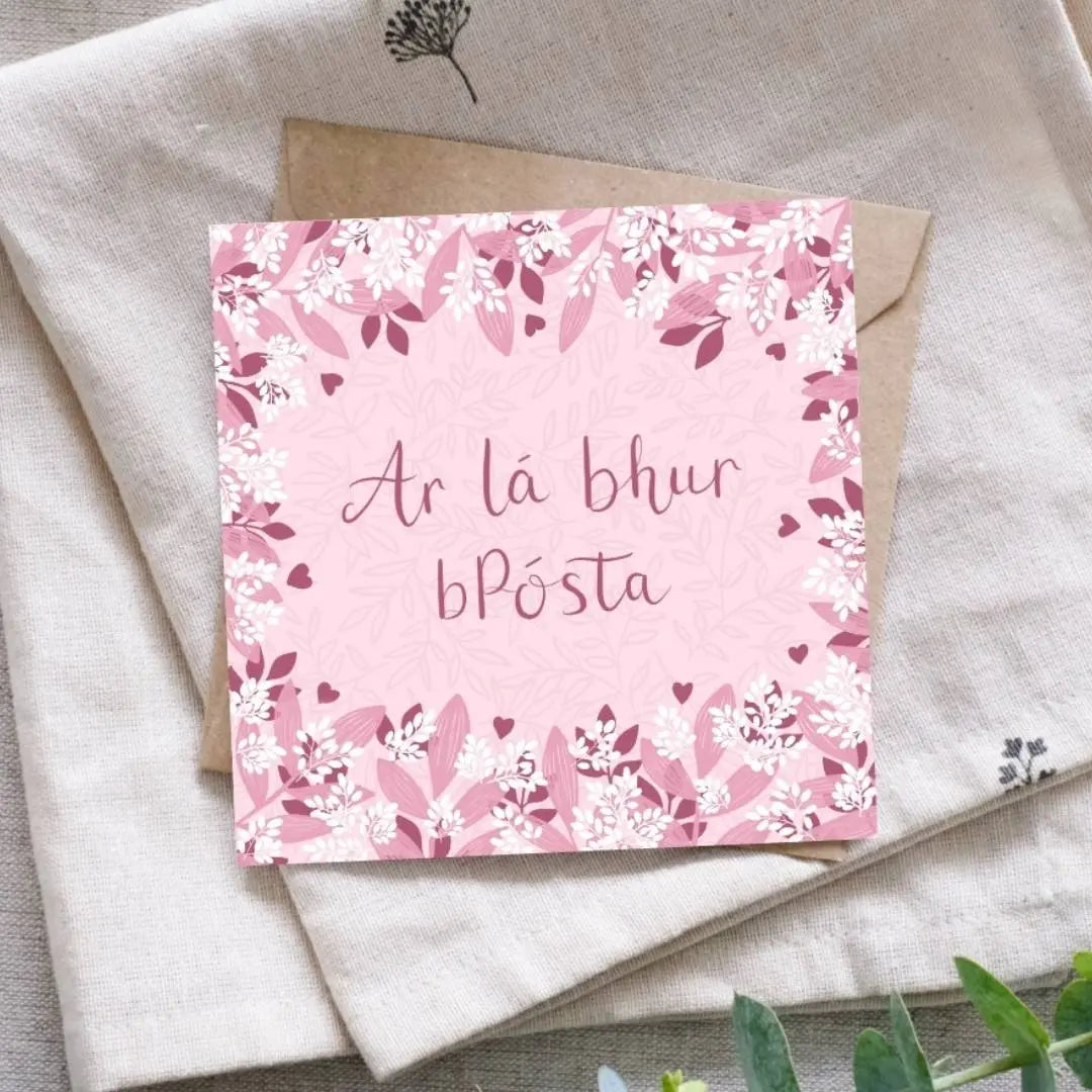 Irish language wedding card with the words "Ar lá bhur bPósta" written in the centre of the card in a dark pink hand writing. Surrounding the writing is a mixture of light and dark pink leaves of various shapes and sizes, as well as sprigs of small white leaf clusters, reminiscent of confetti. Behind the card is a recycled brown Kraft envelope. These are both sitting on a piece of folded cloth with some greenery nearby.