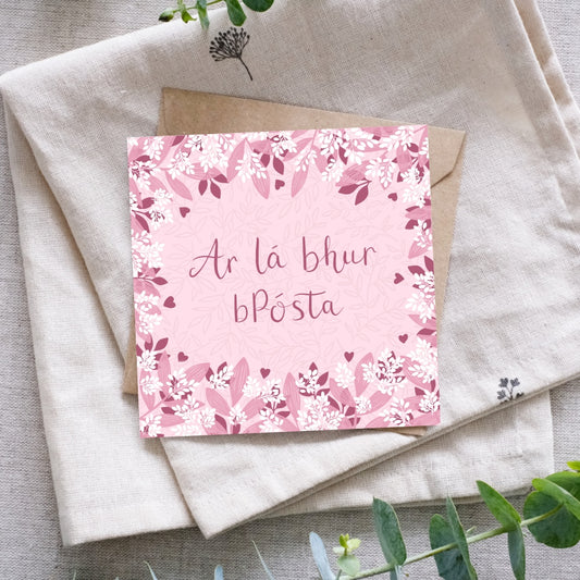 Irish language wedding card with the words "Ar lá bhur bPósta" written in the centre of the card in a dark pink hand writing. Surrounding the writing is a mixture of light and dark pink leaves of various shapes and sizes, as well as sprigs of small white leaf clusters, reminiscent of confetti. Behind the card is a recycled brown Kraft envelope. These are both sitting on a piece of folded cloth with some greenery nearby.