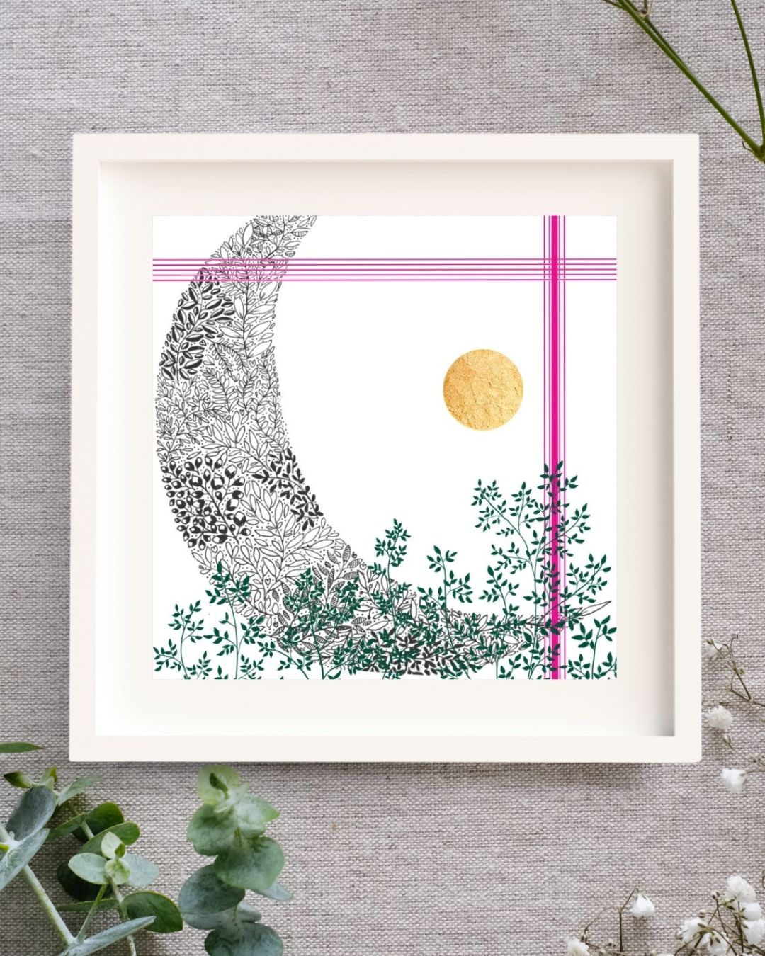 Image shows a framed art print on a grey/brown linen cloth with eucalyptus and baby's breath around it. The frame is white. It's square. There is a white mount surround the print. The print is of a black pen illustration of a moon, blended with some green leaves to the bottom of the print. To the top and the right side there are a few bright pink lines and lastly, to the right of centre there is a hand-applied gold leaf circle.