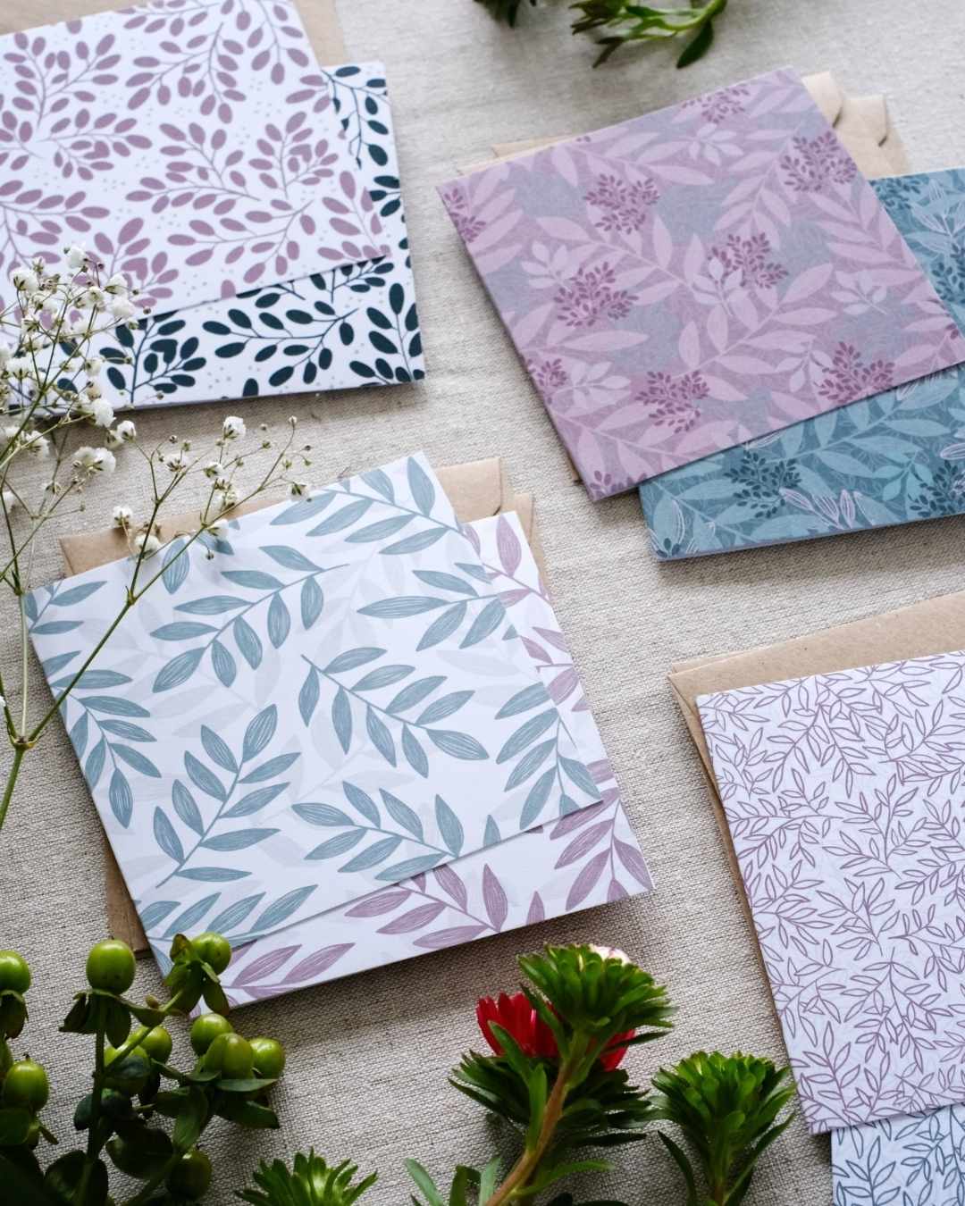 A collection of 8 square botanical-themed note cards, each with their own Kraft brown envelope. The note cards feature intricate leafy patterns in a mixture of green and pink color schemes. The note cards are laid out on a neutral natural fabric surface. Surrounding the card bundles are bunches of greenery, white and pink flowers which go with the natural botanical tone of the sustainably made note cards.