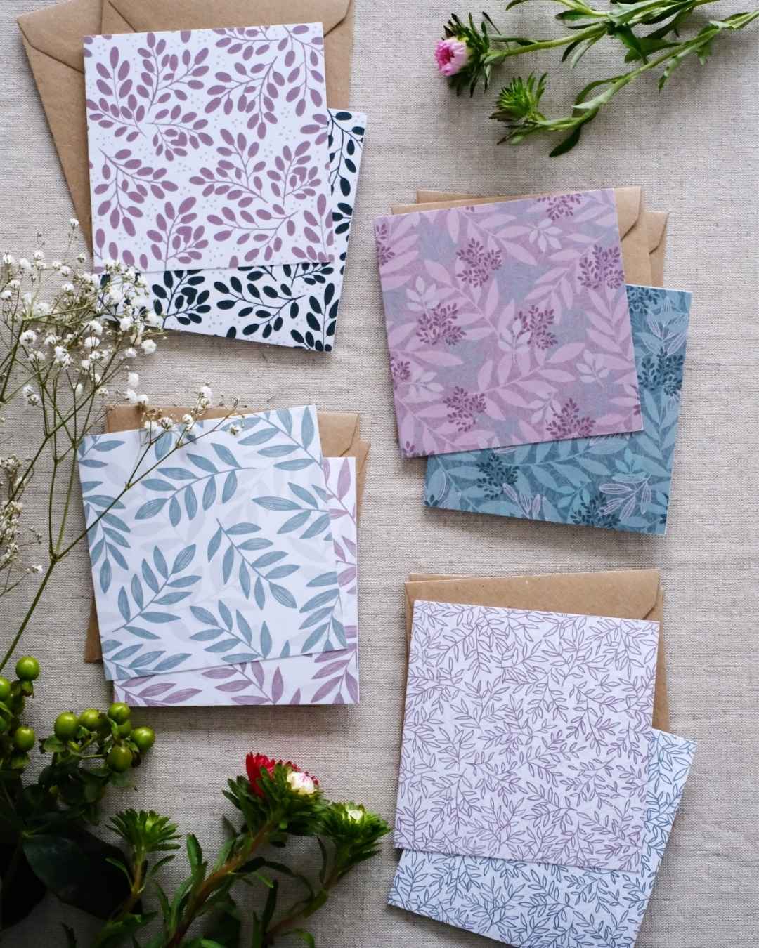 A collection of 8 square botanical-themed note cards, each featuring intricate leafy patterns in green and pink color schemes. The cards are stacked with matching brown kraft envelopes and laid out on a neutral natural fabric surface. Surrounding the card bundles are bunches of greenery, white and pink flowers which go with the natural botanical tone of the note cards.