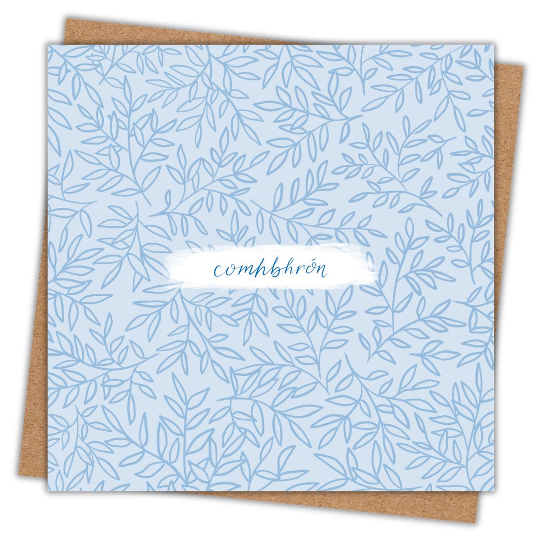 Greeting card with a soft blue leaf pattern on a lighter blue background. In the centre there is a patch on white, on which I have written "Comhbhrón", which is Sympathy as Gaeilge. Behind the card is a brown Kraft envelope.