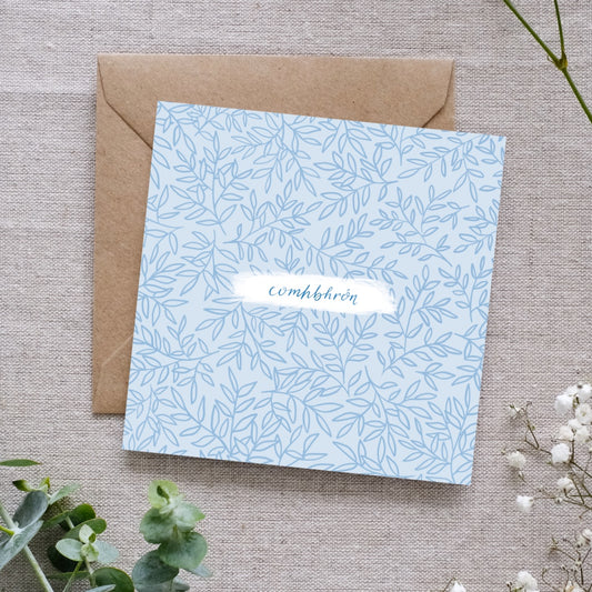 Sympathy card, pale blue with a simple leaf pattern. In the centre it says "Comhbhrón" which means Sympathy in Irish. The writing is my own cursive handwriting in a simple blue. Behind the square card is a brown kraft envelope. They are both laid on a natural cloth with sprigs of foliage and buds around it.