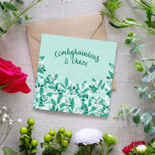 A greeting card with a green and white foliage design, text 'Comhghairdeas Ó Chroí' which is the Irish for 'Heartfelt Congratulations', and an envelope, presented on a background with floral arrangement.