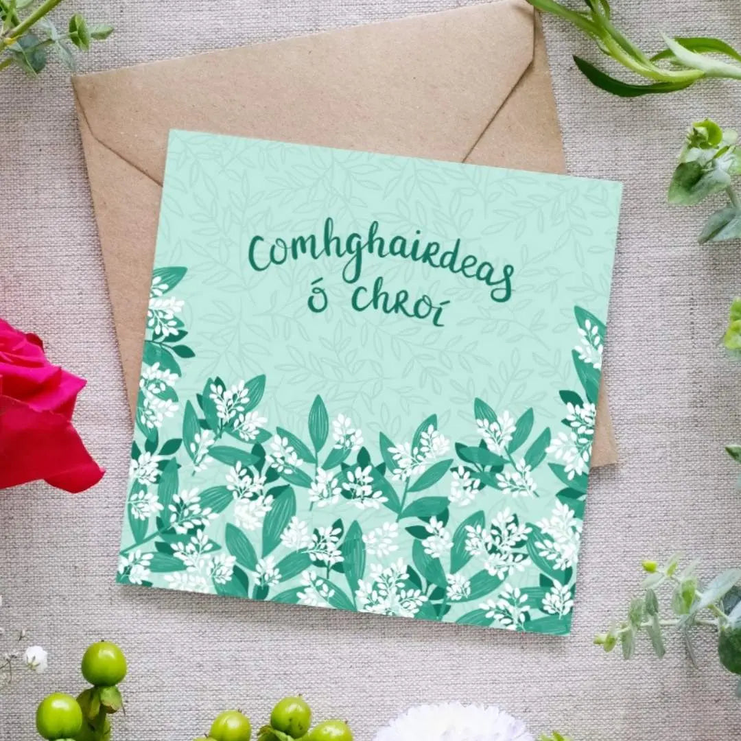 A greeting card with a green and white foliage design, text 'Comhghairdeas Ó Chroí' which is the Irish for 'Heartfelt Congratulations', and an envelope, presented on a background with floral arrangement.