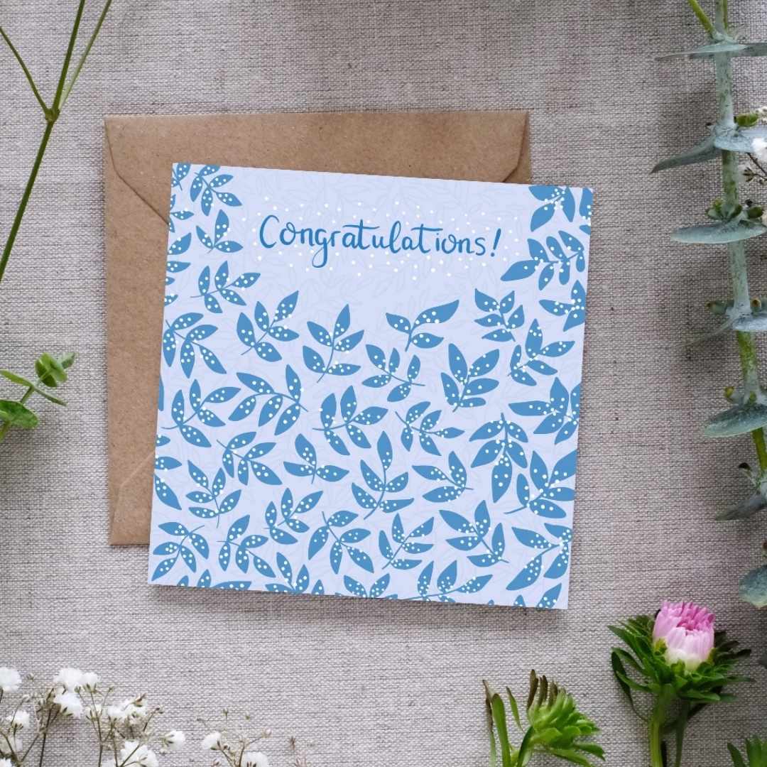 Image shows a congratulations card. The word congratulations is written in dark blue handwriting. In the same dark blue there are clusters of leaves, covered in a white dots which are reminiscent of confetti. The card is sitting on a brown Kraft envelope. Both are laid out on a linen cloth surrounded by light delicate pretty flowers and leaves.