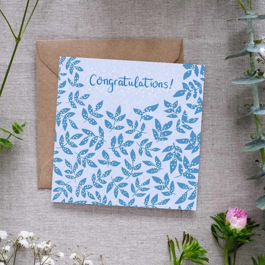 Image shows a congratulations card. The word congratulations is written in dark blue handwriting. In the same dark blue there are clusters of leaves, covered in a white dots which are reminiscent of confetti. The card is sitting on a brown Kraft envelope. Both are laid out on a linen cloth surrounded by light delicate pretty flowers and leaves.