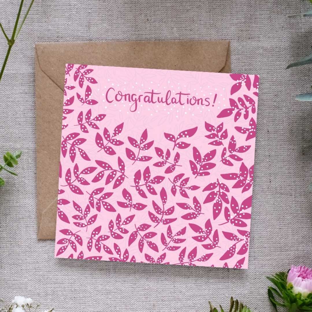 Image shows a congratulations card. The word congratulations is written in deep Pink handwriting with white dots around it. In the same deep pink there are clusters of leaves, covered in a white dots which are reminiscent of confetti. The card is sitting on a brown Kraft envelope. Both are laid out on a linen cloth surrounded by light delicate pretty flowers and leaves.