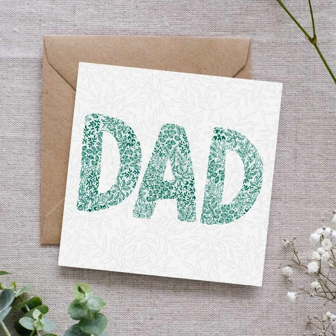 image shows a greeting card with a soft leafy pattern in the background. In the foreground the word DAD is written using intricate leafy details in a beautiful deep green. The card is laid on a brown Kraft envelope. In the background there is a linen cloth and delicate botanical elements.