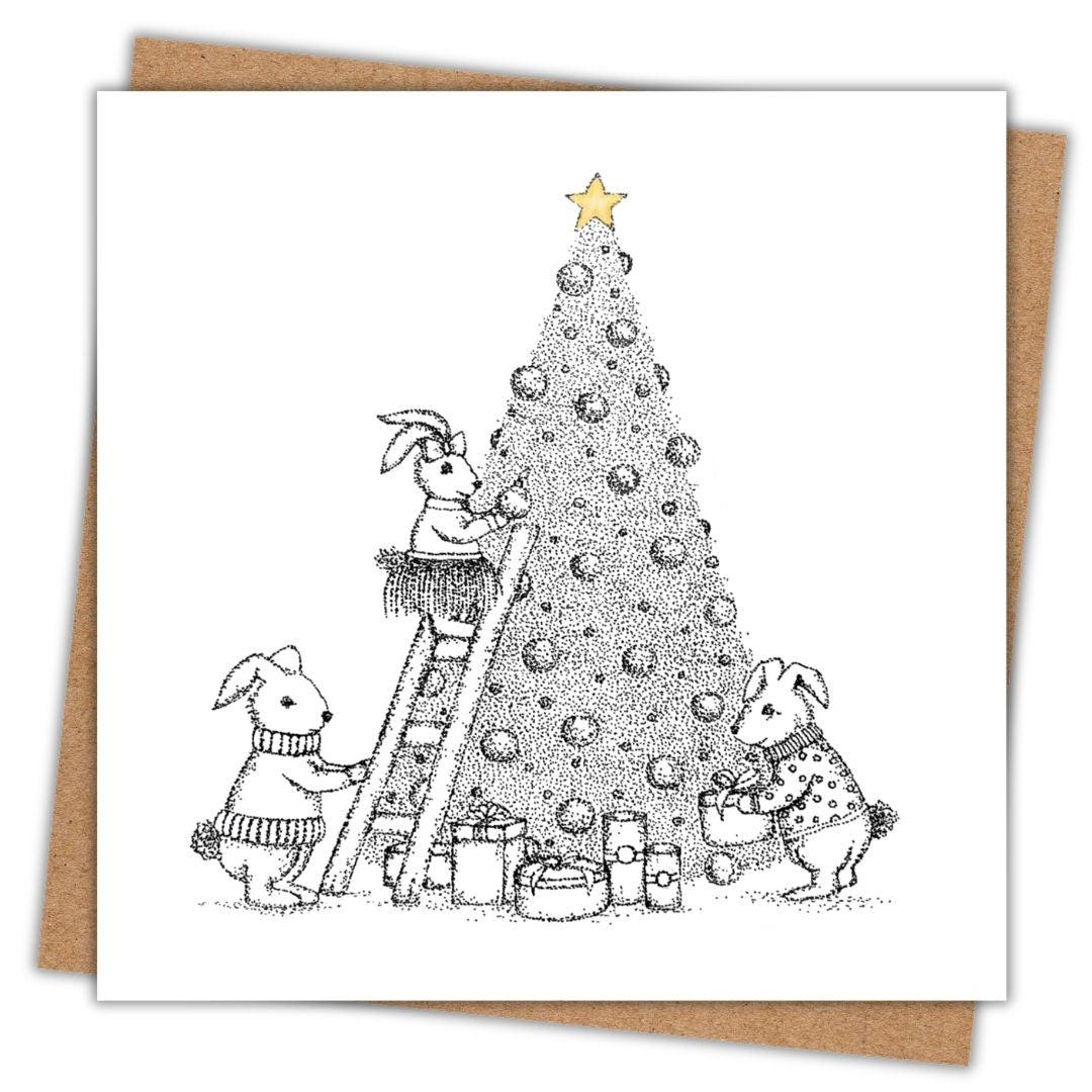 Christmas card of three little bunnies in their festive clothes, decorating a tree in their home with presents underneath and a gold star at the top. The rest of the illustration is black and white.