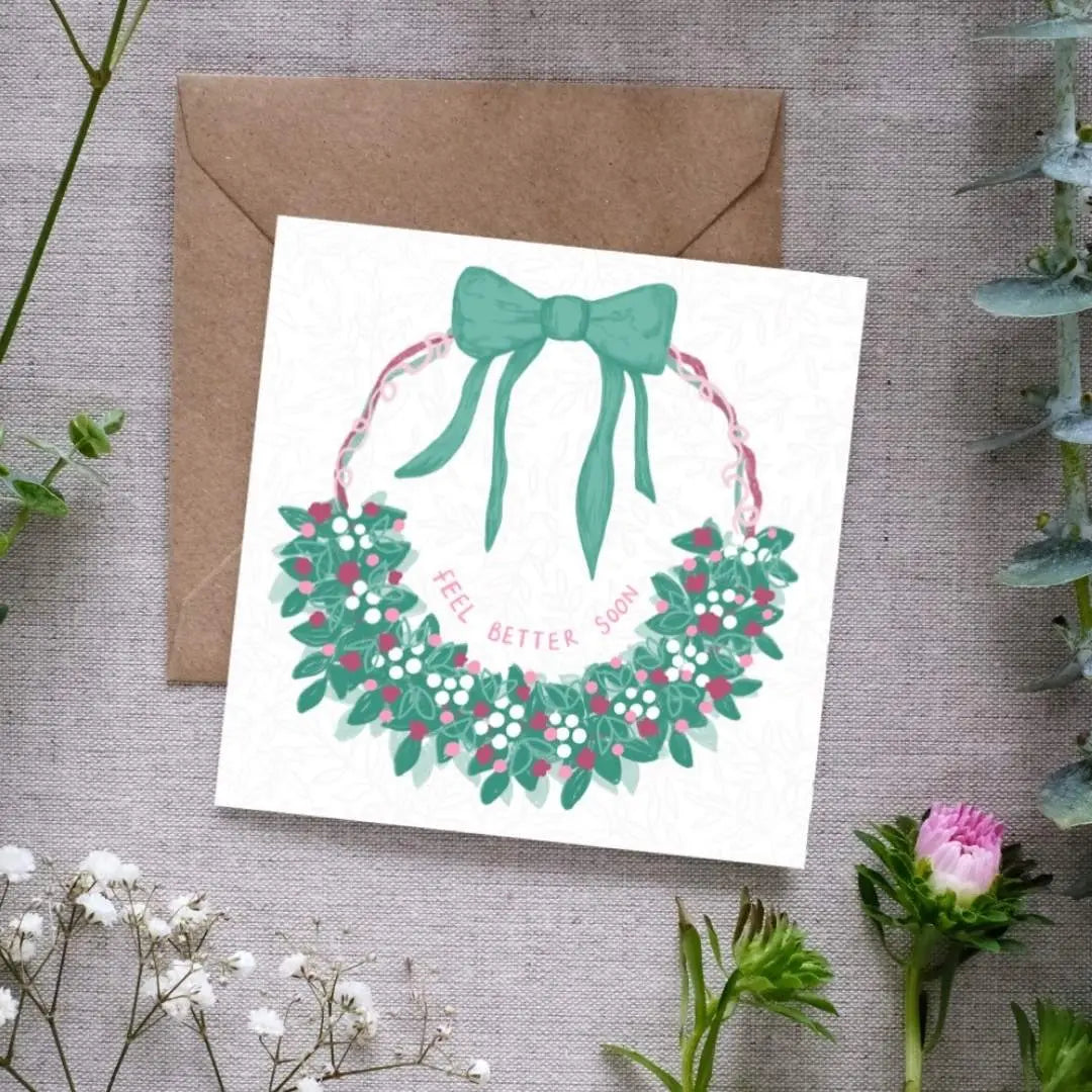 A botanical greeting card with a hand-drawn floral half-wreath on a circle of pink green and purple ribbons leading up to a big green bow. Inside the wreath is a hand-written note that reads 'Feel Better Soon'. The card is accompanied by a brown envelope and placed on a background with floral elements.