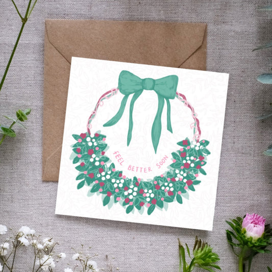 A botanical greeting card with a hand-drawn floral half-wreath on a circle of pink green and purple ribbons leading up to a big green bow. Inside the wreath is a hand-written note that reads 'Feel Better Soon'. The card is accompanied by a brown envelope and placed on a background with floral elements.