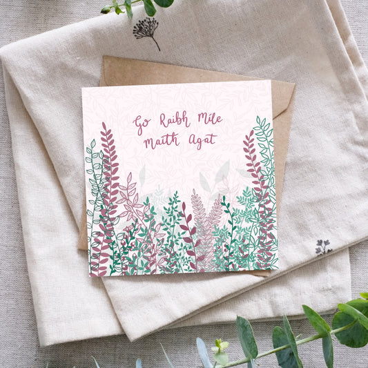 And Irish language thank you card that says "Go Raibh Míle Maith Agat" in Irish along the top of the card. Below the writing are bunches and branches of leaves, and buds in a mixture of dark pink and lots of green tones. Beneath the card is a recycled brown Kraft envelope and they are both sitting on a folded cloth surrounded by foliage for display.
